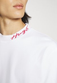 Bílá mikina s kulatým výstřihem a červenou výšivkou "Hug" na límci. Materiál vypadá hladce a má jemnou texturu.