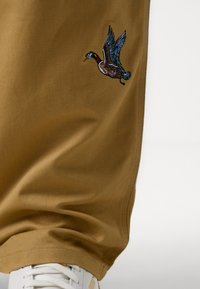 Braune Jogginghosen aus Textil mit glatter Oberflächenstruktur, versehen mit einem gestickten fliegenden Entenmotiv auf dem linken Oberschenkel, kombiniert mit weißen Sneakers.