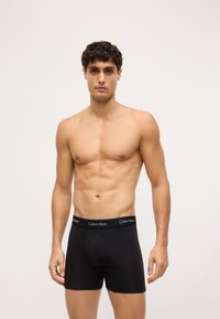 Čierne bavlnené boxerky s priliehavým dizajnom, obsahujú elastický pás s logom a hladkú textúru. Model stojí vzpriamene.