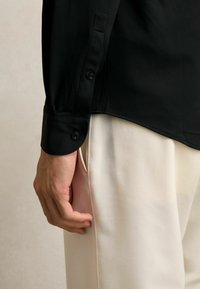 Musta pitkähihaine paita, jossa on sileä pinta ja nappikiinnitys, yhdistettynä vaaleisiin housuihin, esittäen siistiä ja minimalistista muotoilua.