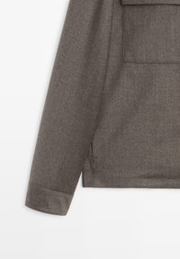 Tessuto marrone testurizzato con un design sartoriale. Presenta una tasca, una manica con polsino e un orlo pulito. Finitura opaca, senza ulteriori abbellimenti.