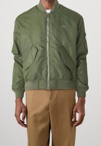 Bomber bunda v olivově zelené barvě s předním zipem, žebrovanými manžetami a dvěma kapsami s klopami. Má malý logo na hrudi a hladkou texturu.