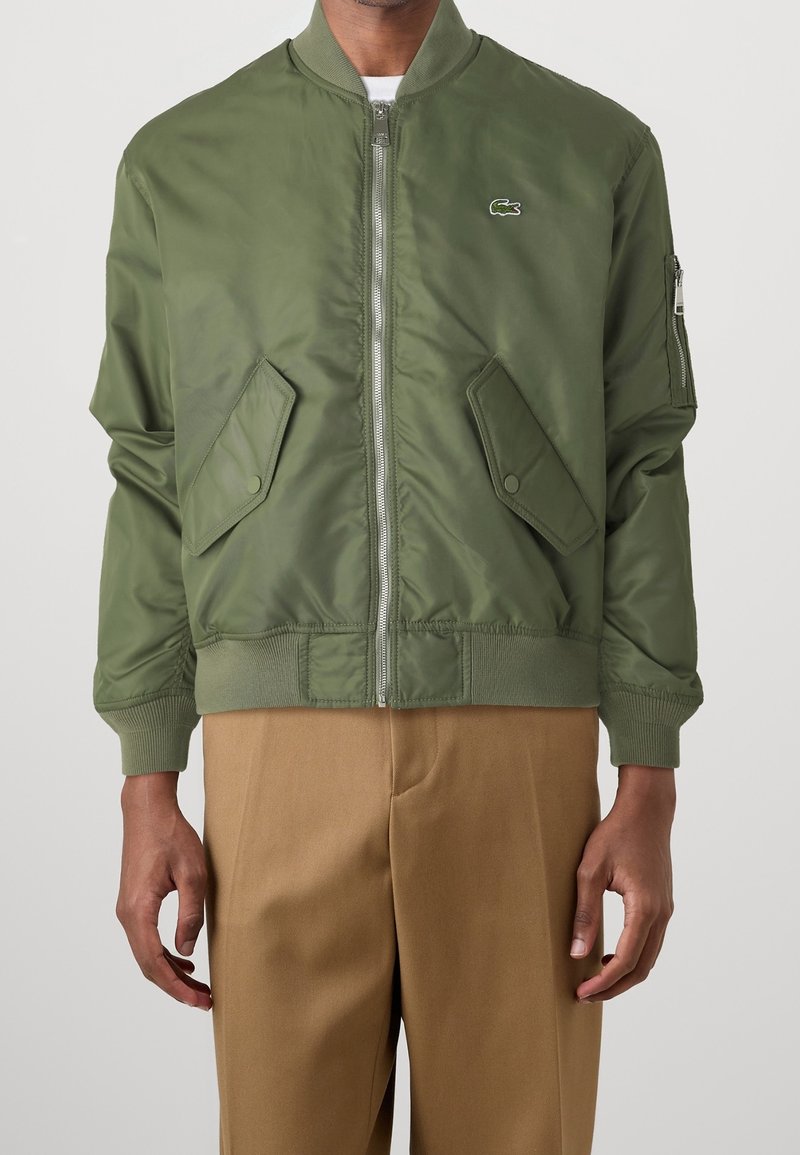 Bomber bunda v olivově zelené barvě s předním zipem, žebrovanými manžetami a dvěma kapsami s klopami. Má malý logo na hrudi a hladkou texturu.