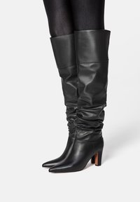 Black leather knee-high boots with a slouchy design, pointed toe, and chunky heel. Smooth texture with a shiny finish and folded top.