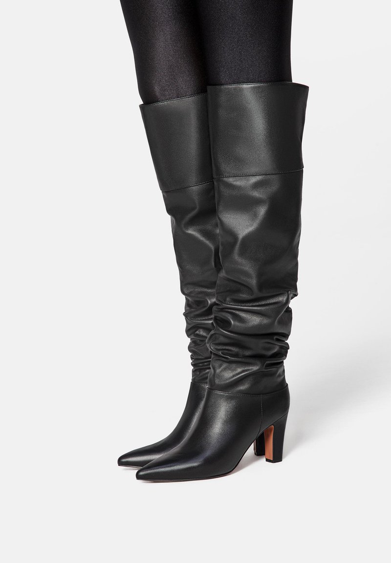 Black leather knee-high boots with a slouchy design, pointed toe, and chunky heel. Smooth texture with a shiny finish and folded top.