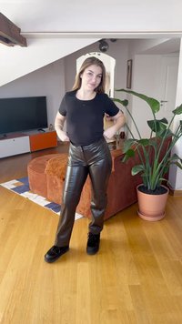 Juoda prigludusi marškinėliai ir aukšto liemens blizgios rudos netikros odos kelnės. Modelis stovi ant medinių grindų ryškioje patalpoje su augalais.