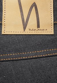 Denimtyg med en läderlapp. Lappen har en svart "W"-logotyp och "Nudie Jeans Co" i liten text. Kontrastfärgad orange sömnad längs kanterna.