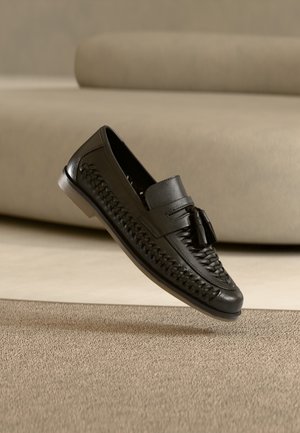 Mocasín tejido de cuero negro con borlas, descansando con la suela inclinada hacia arriba sobre una alfombra beige frente a un fondo de sofá beige.