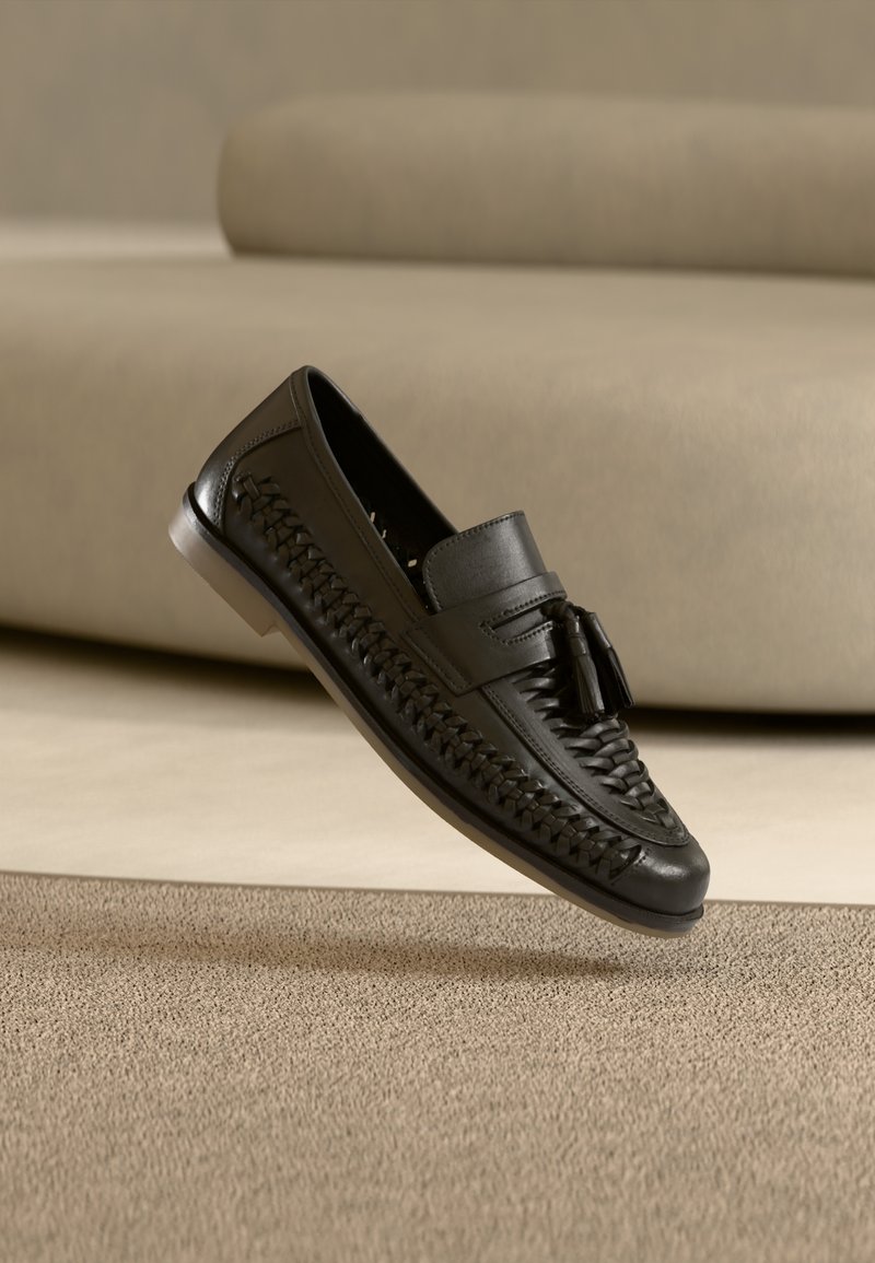 Czarna skórzana pleciona mokasyna z frędzlami, spoczywająca pod kątem podeszwą do góry na beżowym dywanie przed beżową sofą.