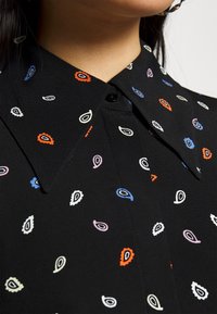 Blusa preta com colarinho e padrões em forma de gota de várias cores, incluindo vermelho, azul, branco e rosa. Tecido suave com textura sutil e fecho de botão.