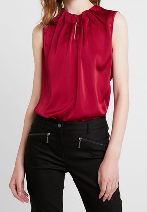 Ærmeløs rød bluse med rynket halsudskæring og lille nøglehuldsdetalje, kombineret med sorte bukser med vandrette lynlåslommer foran.