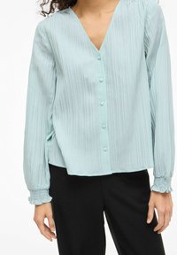 Gaismi zila, garas piedurknes blūze ar vertikālām svītrām, V veida kakla izgriezumu, pogām priekšpusē un savilktām aprocēm. Brīvs piegriezums, savienots ar melnām biksēm.