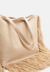 Sac fourre-tout tissé beige avec franges à la base, doté de anses robustes et texturées ainsi que d'un design à ouverture supérieure pour un accès facile.