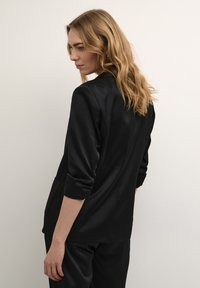 Blazer in satin nero con un design su misura, con maniche arrotolate e tasche laterali, mostrato di spalle su uno sfondo neutro.