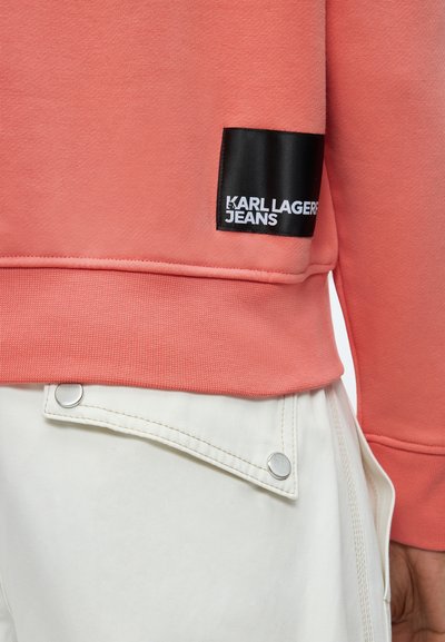 Макро на подгъва на коралова блуза със черен етикет „Karl Lagerfeld Jeans“ над бели панталони със сребристи копчета.