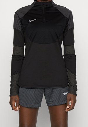 Person trägt ein langärmeliges Nike-Oberteil in Schwarz und Dunkelgrau mit Viertelreißverschluss und graue Nike-Shorts mit Muster.