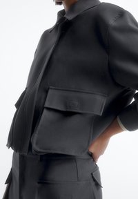 Sort jakke med en frontlynlås, to store klaplommer og en kravehals. Materialet ser glat ud med en mat tekstur.