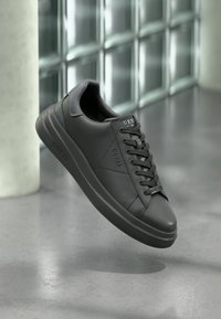Baskets noires de la marque Guess avec lacets flottant au-dessus d'un sol en béton poli devant un mur en blocs de verre.