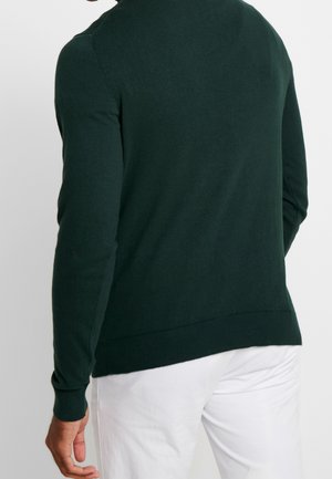 Homme portant un pull vert foncé à manches longues et un pantalon blanc, vu de dos sur un fond clair uni.