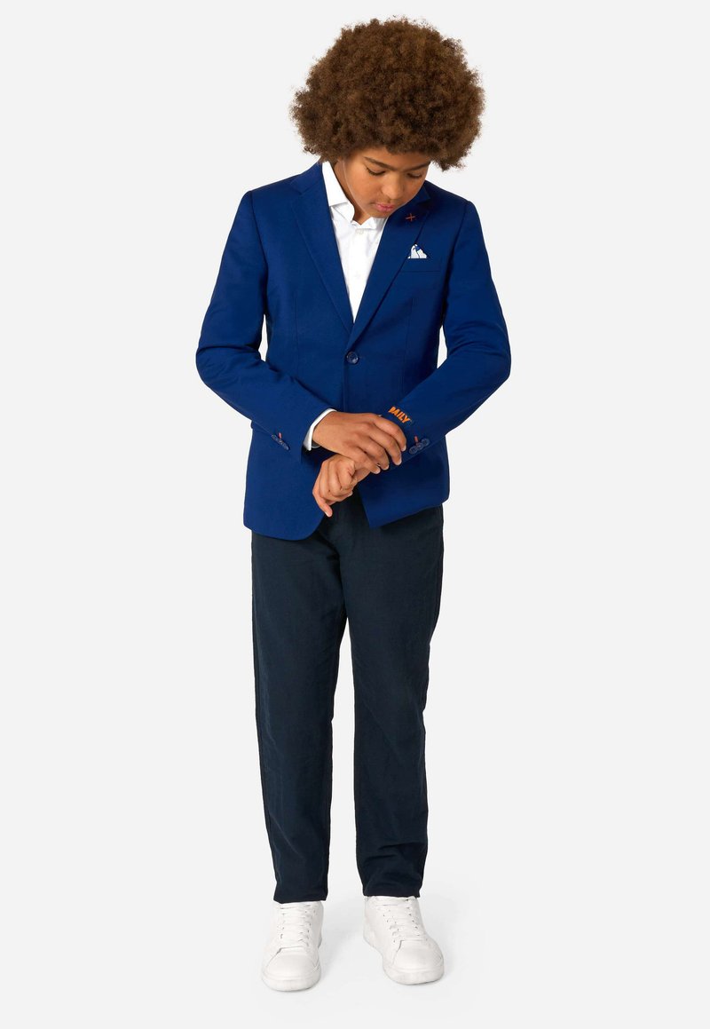 Zils žakets ar pielāgotu griezumu, balta krekls, tumši bikses un baltas kedas. Iekļauj oranžu izšuvumu uz piedurknes un dekoratīvas pogas.