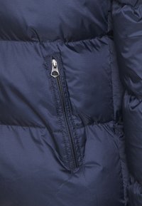 Marinblå puffjacka i tyg med kviltade sektioner, med en metallisk dragkedjeficka och en slät, strukturerad yta.