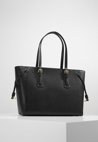 Bolso tote de cuero negro texturizado con doble asa, acentos de hebilla dorada y correas laterales ajustables, exhibido en un pedestal blanco.