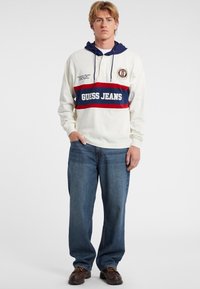 Witte hoodie met marineblauwe accenten, een rode en marineblauwe streep, en de tekst "GUESS JEANS". Eenvoudig te combineren met blauwe jeans en bruine schoenen. Casual pasvorm.