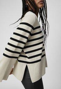 Cremefarvet sweater med sorte vandrette striber, ribbet tekstur, flared ærmer, slidser i siderne og en afslappet silhuet.