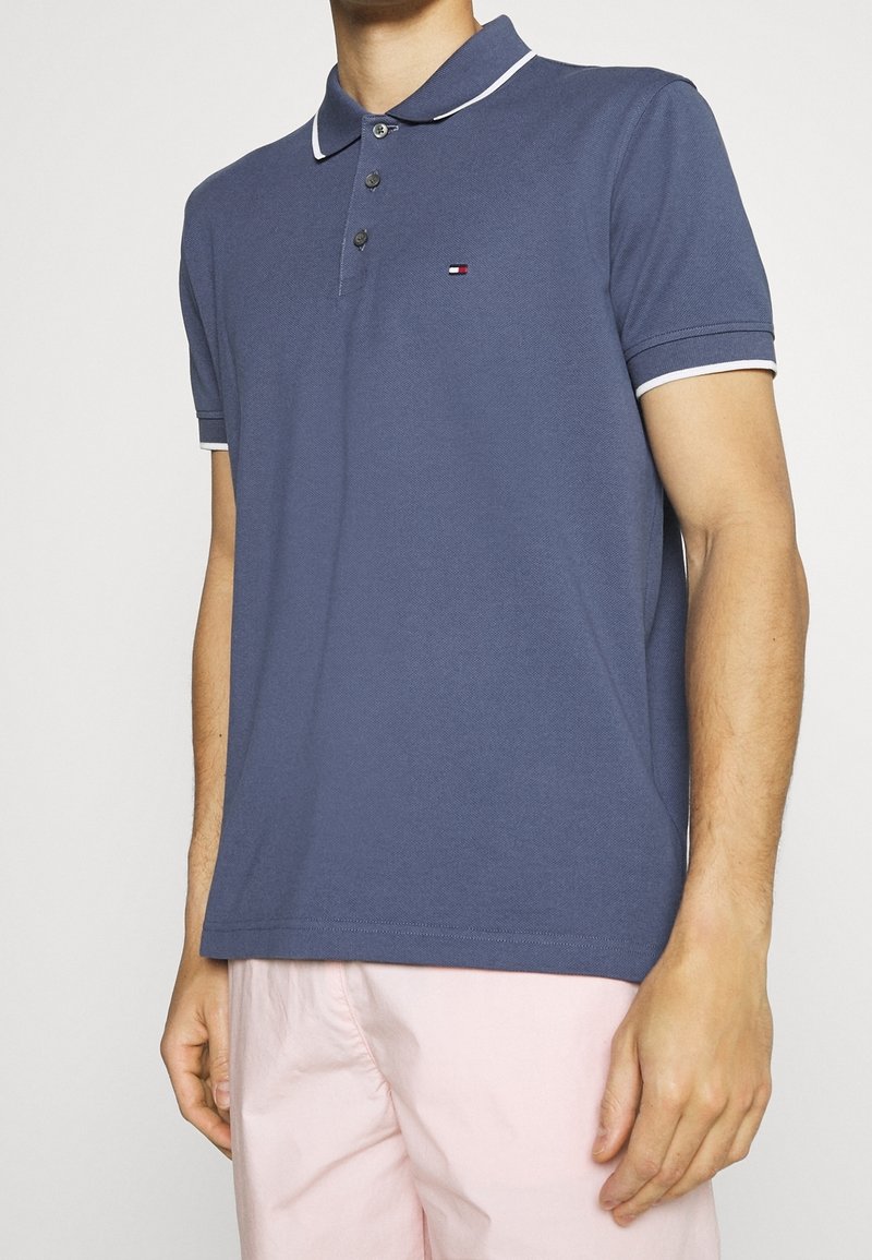 Homme portant un polo à manches courtes bleu avec des bordures blanches et un pantalon rose pâle, se tenant devant un fond uni.
