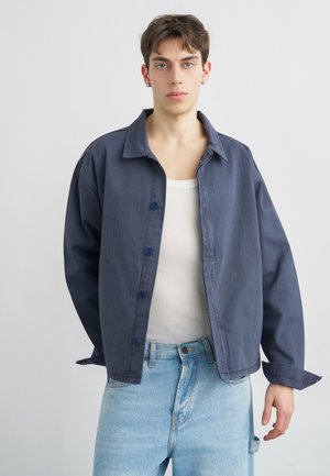 Jonge persoon die een losse donkerblauwe jas met knopen draagt, een wit geribd tanktopje en lichtblauwe jeans, staand tegen een effen achtergrond.