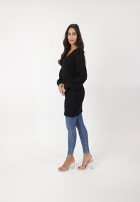 Pull en tricot noir oversized à manches longues, associé à un jean skinny bleu et des sandales à talons bloc transparent sur un fond blanc.