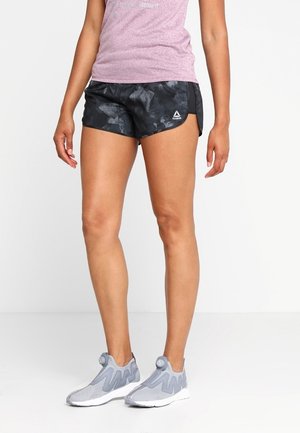 Kvinde iført sorte mønstrede Reebok-shorts, en pink sportstrøje og grå slip-on løbesko, stående mod en hvid baggrund.