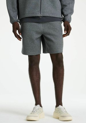 Mann trägt graue Shorts, passende Reißverschlussjacke und weiße Turnschuhe, steht vor einem schlichten weißen Hintergrund.