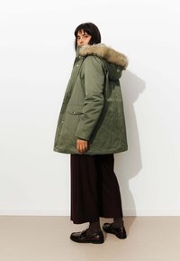 Parka olive verte avec capuche en fourrure, boutons pression, poches latérales et coupe ample, associée à un pantalon large sombre et des chaussures.