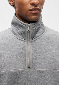 Γκρι φούτερ με φερμουάρ που διαθέτει ψηλό κολάρο, μαλακή υφή και αντίθετη ραφή κατά μήκους των ραφών και του φερμουάρ.