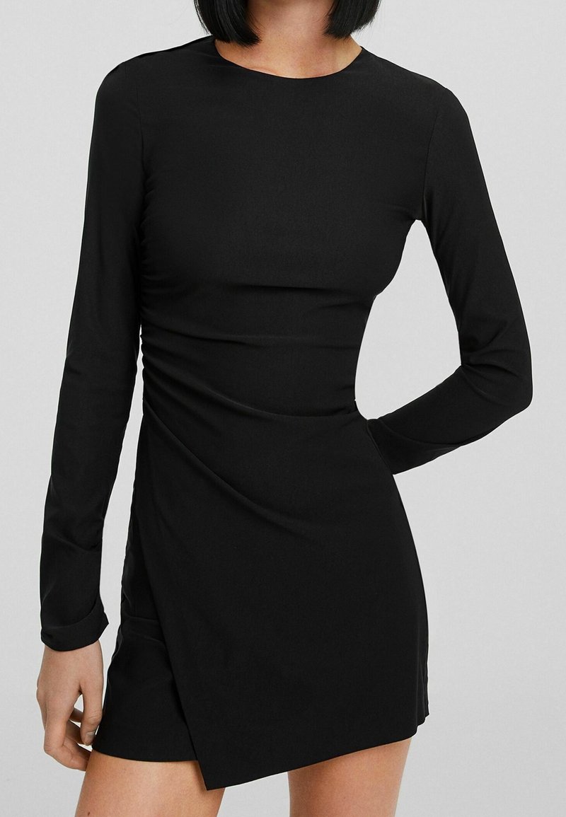 Femme portant une robe mini noire ajustée à manches longues avec un ourlet asymétrique et un bras plié derrière le dos.