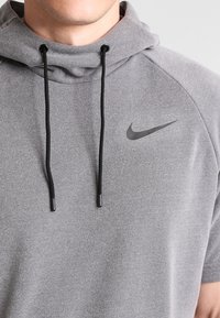 Sweat-shirt à capuche gris en tissu texturé, avec une capuche, des cordons de serrage noirs, et un petit logo Nike noir sur la poitrine. Manches courtes.