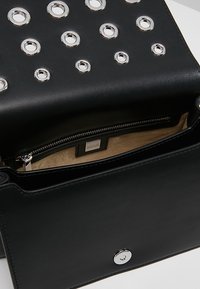 Bolso de cuero negro con detalles de ojales plateados, bolsillo con cremallera en el interior y cierre magnético. Forro suave de color canela visible. Forma rectangular simple.