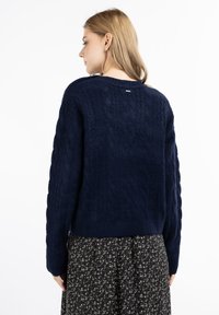 Pulover tricotat cu model din cabluri, de culoare bleumarin, cu guler rotund, corp texturat și tiv cu nervuri. Are o etichetă mică argintie pe spate.