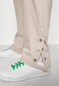 Béžové kalhoty s kovovými tlačítky na lemu, spárované s bílými sneakers s zelenými tkaničkami a tečkovanou podrážkou.