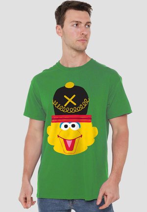 Mann in grünem Shirt mit dem Gesicht eines gelben Cartoon-Vogels, dazu einen schwarzen Hut mit einem gelben Schwirl und roten Streifen.