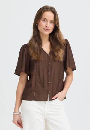 Femme aux longs cheveux bruns portant un chemisier marron boutonné et un pantalon blanc, debout avec une main dans la poche sur un fond uni.