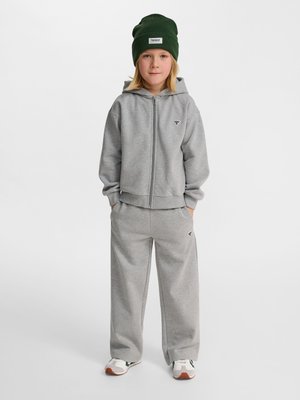 Barn står med front mod kamera iført en grå hættetrøje med lynlås, matchende bukser med vide ben, hvide sneakers og en mørkegrøn hue. Hænderne i lommerne.