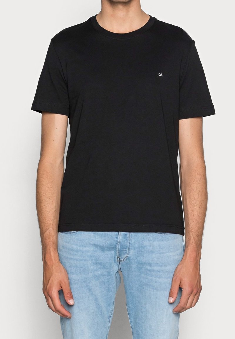 Mężczyzna w prostym czarnym t-shircie z krótkim rękawem z małym białym logo "ck" na piersi i jasnoniebieskich dżinsach, stojący na tle białym.
