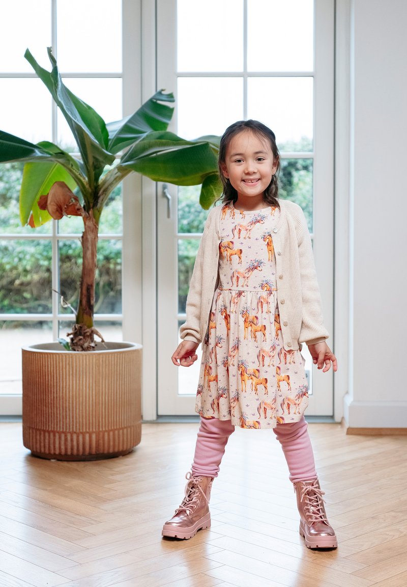 Ein Kind trägt ein beiges Kleid mit Pferdemustern und eine cremefarbene Strickjacke, pinke Leggings und glitzernde rosa Stiefel und steht in einem hellen Raum.
