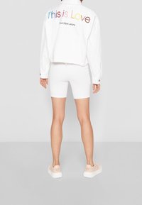 Valkoinen lyhyt farkkutakki, jossa on kirjailtuna monivärisesti "Tämä on rakkautta", yhdistettynä valkoisiin tiukkoihin shortseihin ja vaaleanpunaiseen lenkkaripariin.