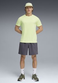 Άνδρας που στέκεται με τα χέρια πίσω από την πλάτη, φοράει ανοιχτοπράσινη μπλούζα Puma, γκρι σορτς, άσπρες κάλτσες Puma, πράσινα αθλητικά παπούτσια και ανοιχτοπράσινο καπέλο.