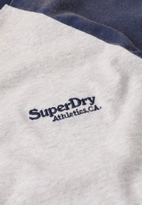 Lichtgrijze en marineblauwe stof met geborduurd "SuperDry Athletics, CA."-logo op lichtgrijze textiel.