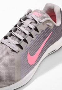 Scarpa da ginnastica grigia con tomaia in rete e logo Nike rosa. Caratterizzata da un design testurizzato, chiusura con lacci e suola in gomma bianca.