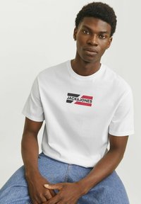 Hvid bomulds-t-shirt med et sort og rødt JACK & JONES logo. Klassisk crew neck design med korte ærmer, parret med blå denim jeans.